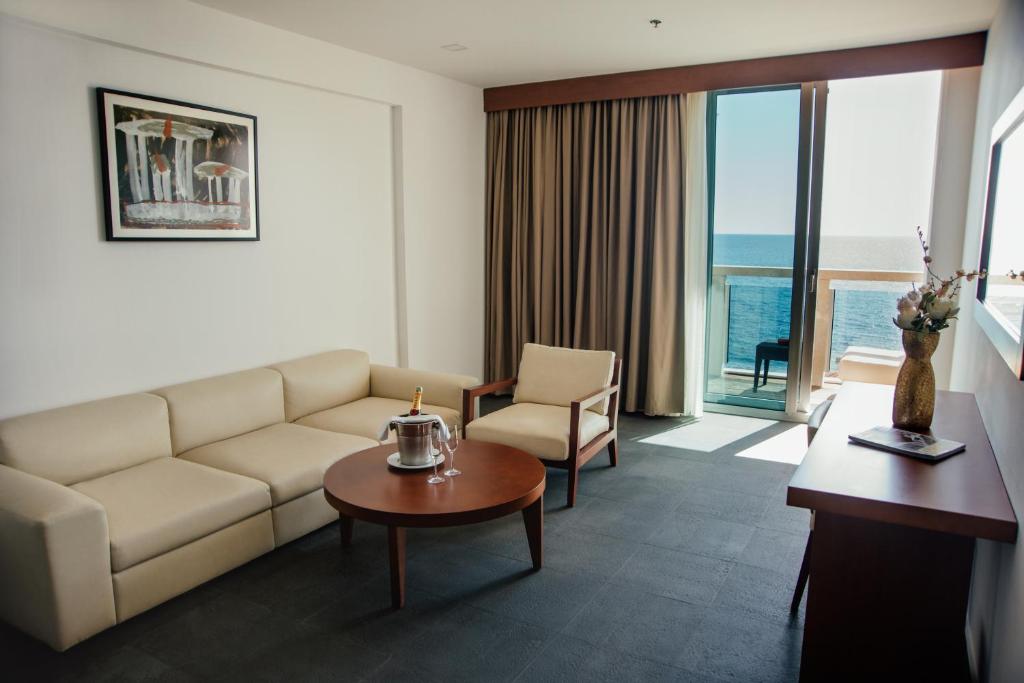 Mūsdienīgs viesnīcas numurs ar jūras skatu, kurā ir ērta dīvāna zona, kafijas galdiņš un liels logs ar balkonu. Sienā ir mākslas darbs, bet blakus stāv galds ar ziediem un dekoriem.