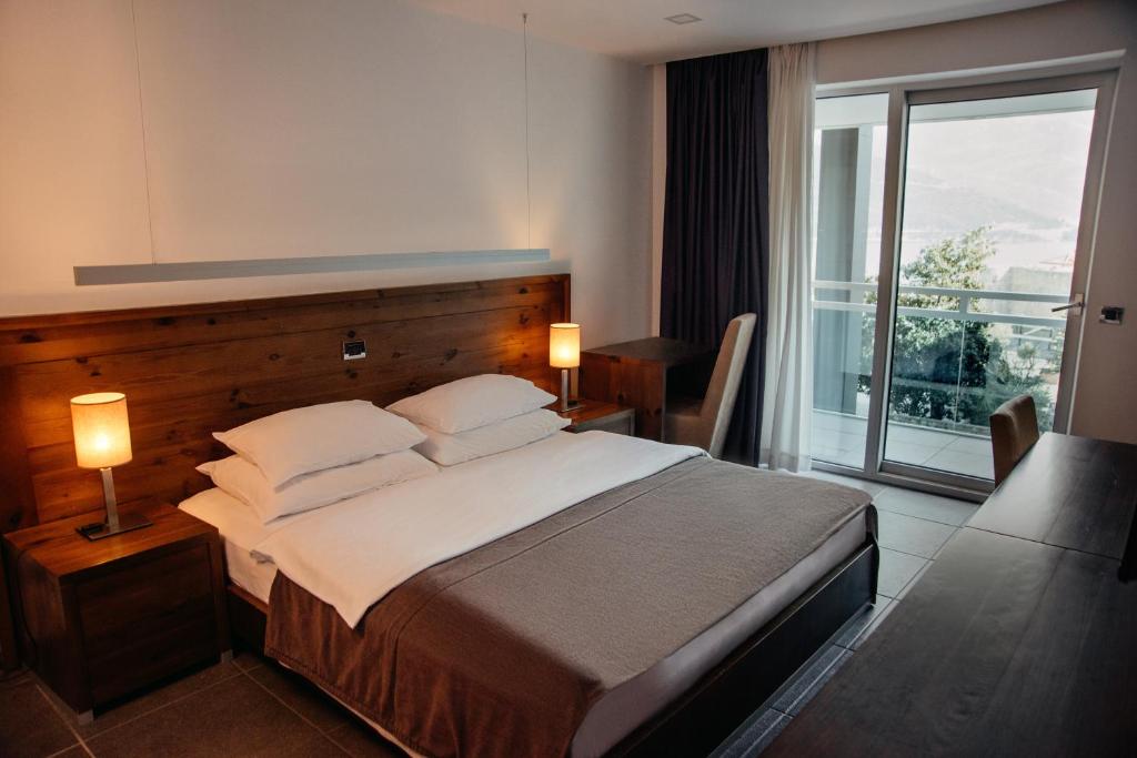 Mūsdienīga viesnīcas numura interjers ar divguļamo gultu, koka elementiem un balkonu, kas nodrošina skatu uz dabu. Apgaismojumu veido stilīgas lampas, radot siltu atmosfēru.