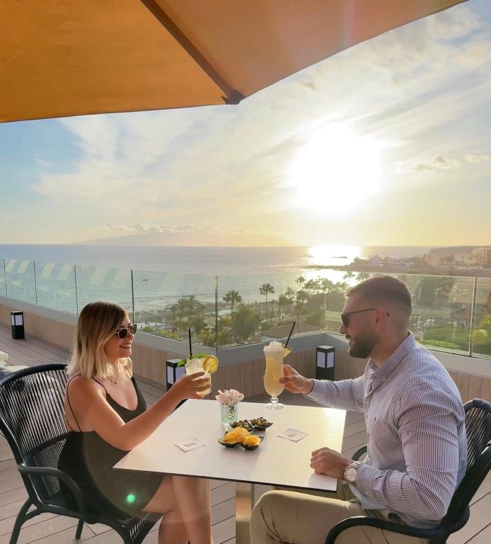 Pāris, izbaudot kokteiļus terasē ar skatu uz saulrietu un okeānu, relaksējas romantiskā atmosfērā.