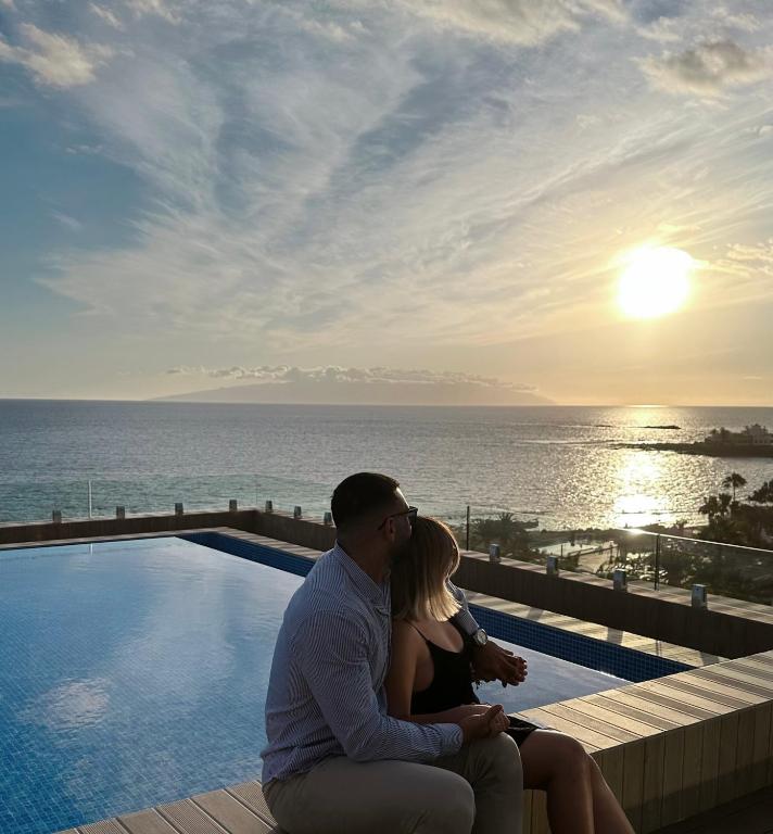 Romantiska ainava ar pāri, kas sēž pie baseina, skatoties uz saulrietu pār jūru. Fons ar zilu ūdeni un debesīm, kuras rotā mākoņi.