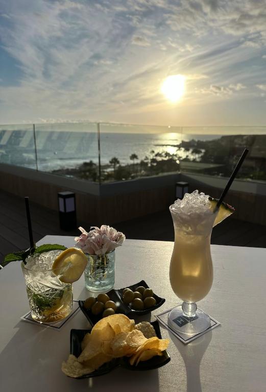 Vasaras kokteiļi un uzkodas ar skatu uz saulrietu pie jūras, radot relaksējošu atmosfēru.