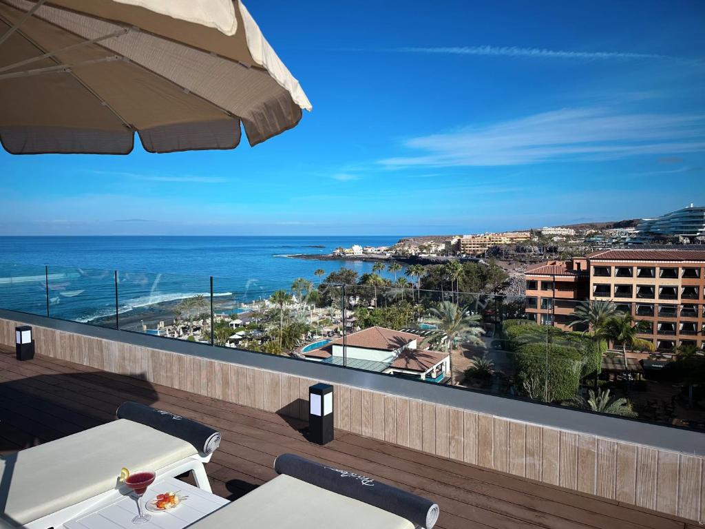 Skats no terases uz skaisto okeānu, ar sauļošanās krēsliem un kokteili uz galda. Aiz tā redzams pludmales komplekss un debesis.