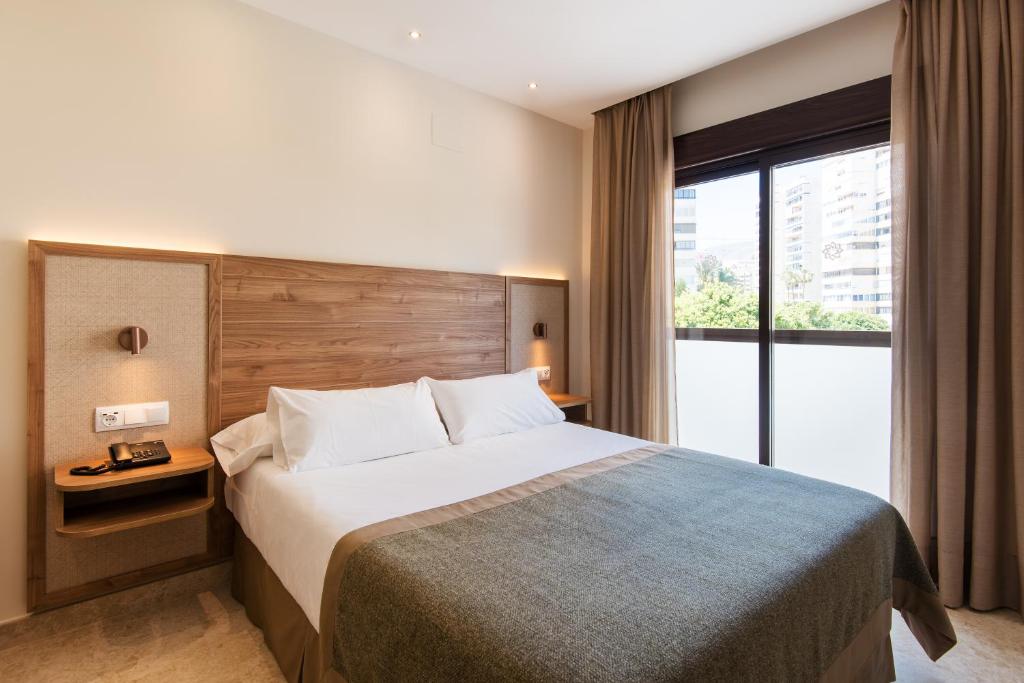 Mūsdienīga viesnīcas istaba ar divguļamo gultu, koka akcentiem un lieliem logiem, caur kuriem ieplūst dabiska gaisma. Elegants un komfortabls interjers rada relaksējošu atmosfēru.