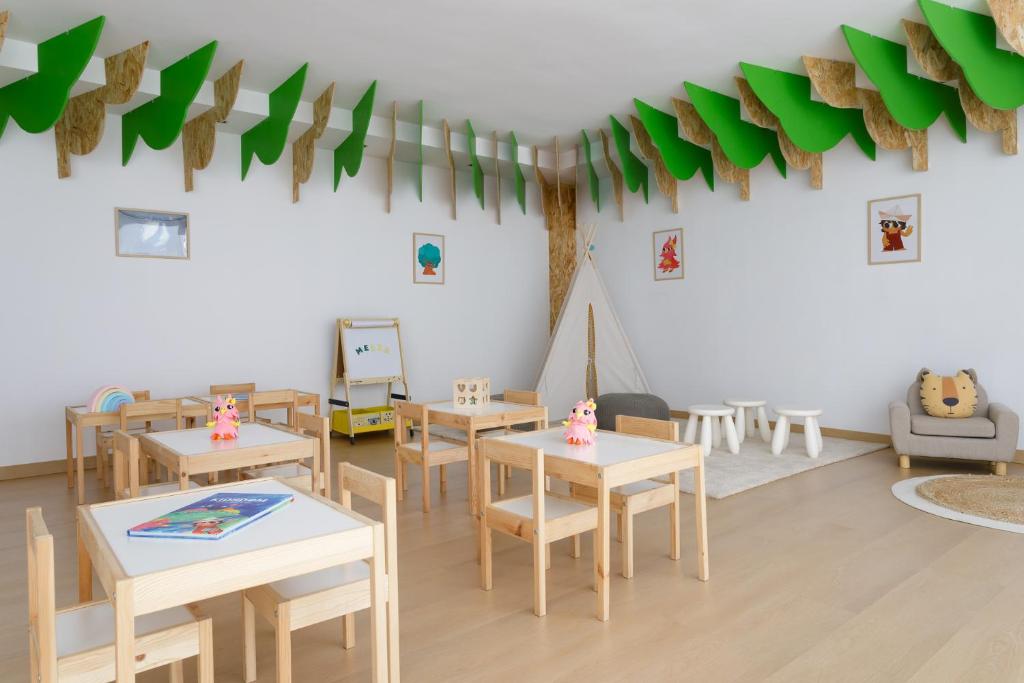 Bērnu rotaļu telpa ar koka galdiem un krēsliem, ko rotā zaļas lapu dekorācijas. Telpā ir rotaļlietas, zīmēšanas dēlis un mīksts paklājs ar ērtu atpūtas stūrīti.