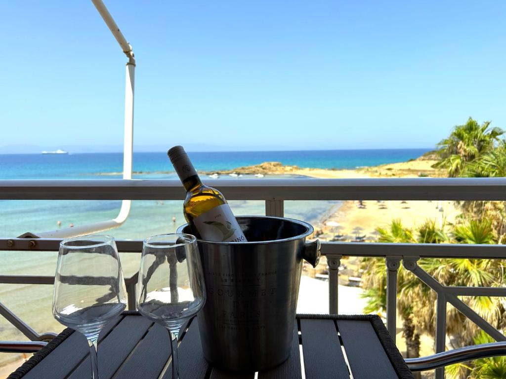 Balto vīna pudele ar divām glāzēm atrodas uz galda ar skatu uz pludmali un jūru. Fons ir saulains, ar palmām un skaidru zilu debesu.