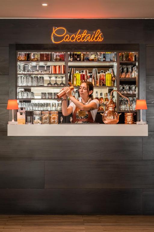 Bāra darbiniece sajauc kokteiļus pie modernā bāra, kurā redzama plaša alkoholisko dzērienu kolekcija. Uz sienas ir izcelts "Cocktails" neona uzraksts.