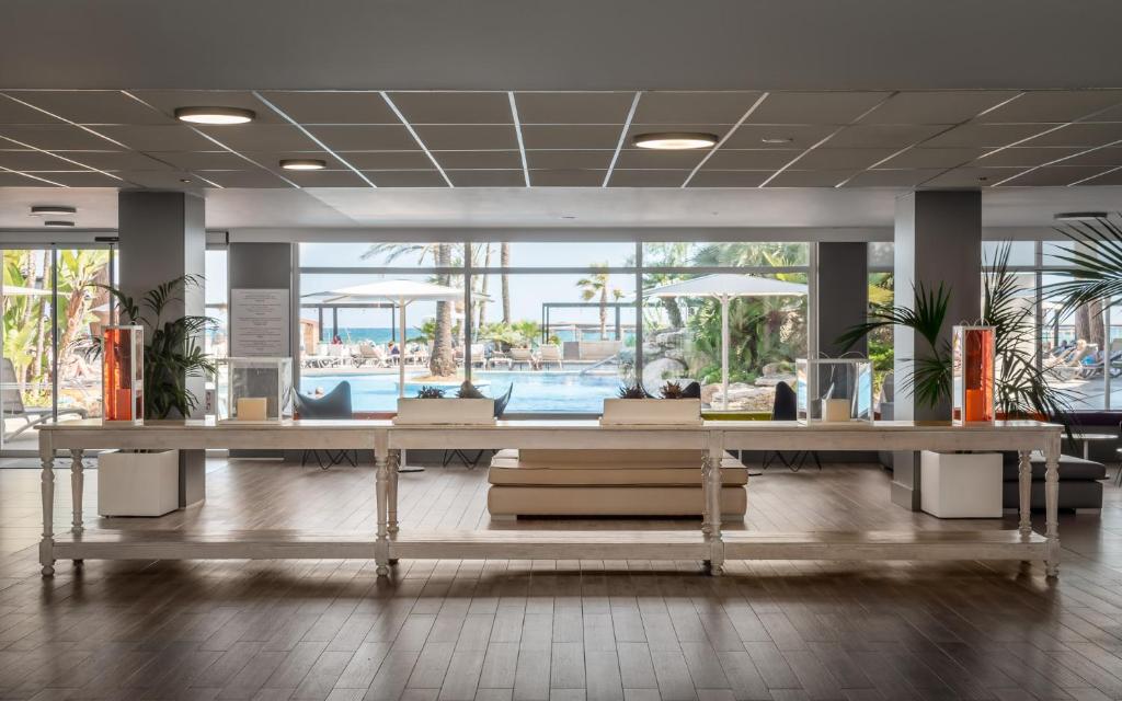Mūsdienīgs viesnīcas reģistratūras interjers ar skatu uz baseinu un palmas, kas rada relaksējošu atmosfēru. Elegantā mēbeļu izkārtojuma un apgaismojuma kombinācija piešķir telpai eleganci.