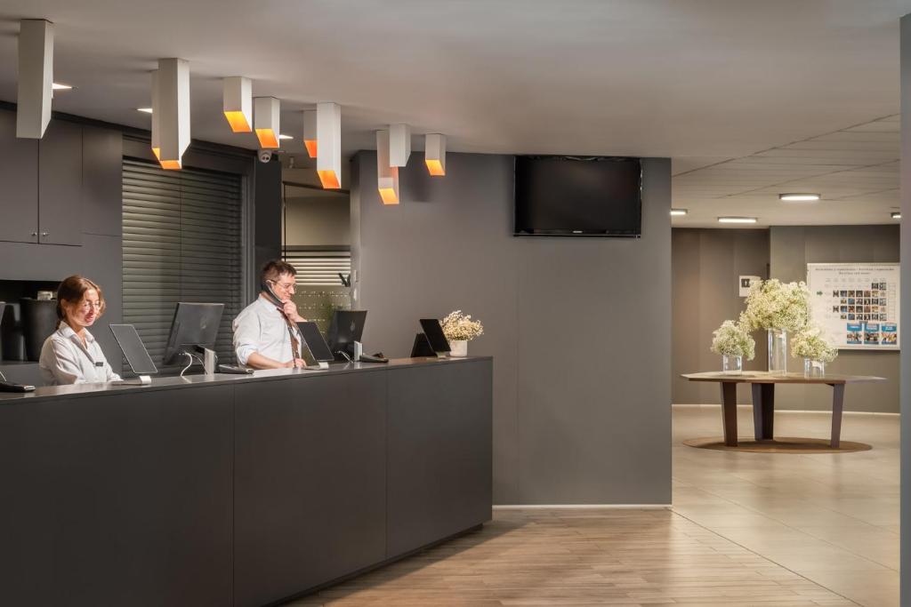 Recepcijas zona ar diviem darbiniekiem, moderns dizains un ziedi uz galda.