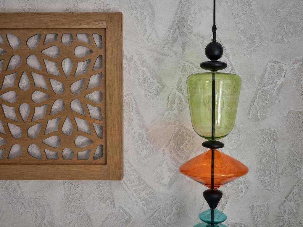 Krāsainas stikla dekoratīvās formas piekārtas pie sienas, blakus koka akcentam ar sarežģītu rakstu.