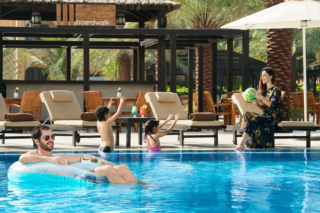 Ģimenes brīvdienas pie baseina, kur pieaugušais relaksējas uz pludiņa, kamēr bērni spēlējas un priecājas pie ūdens. Fons ar palmām un sauļošanās krēsliem rada atpūtas atmosfēru.