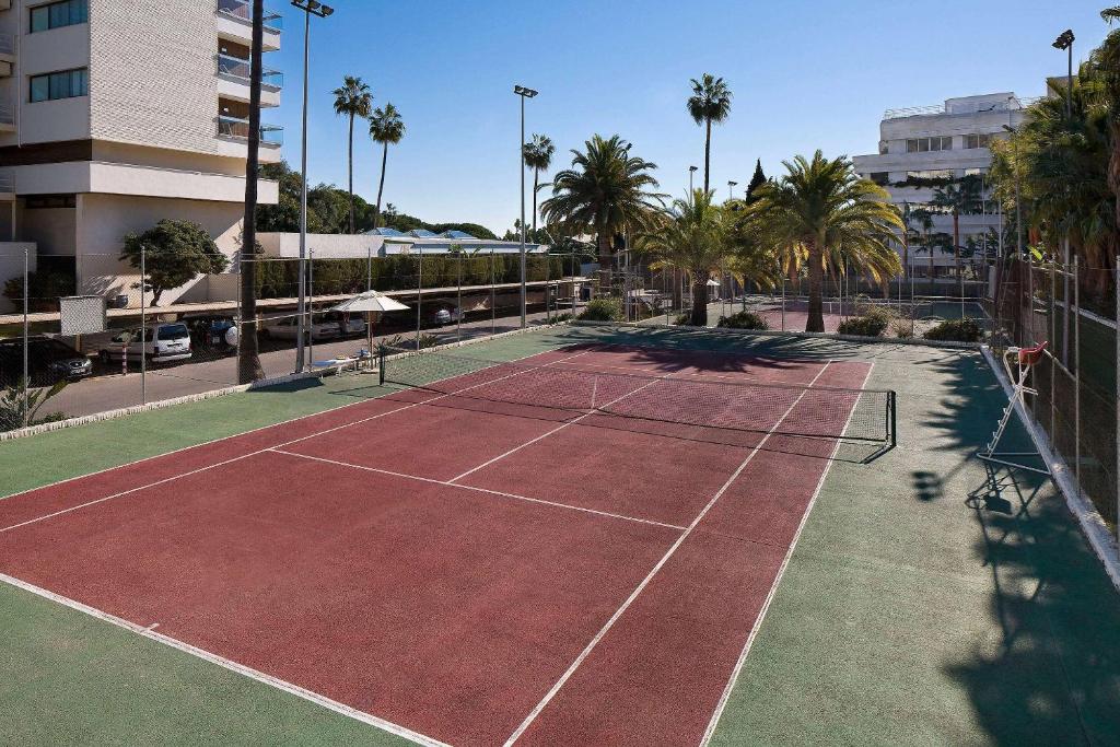 Tennisa laukums ar sarkanu segumu un palmām apkārt, kas atrodas saulainā vidē. Ideāla vieta sportiskām aktivitātēm.
