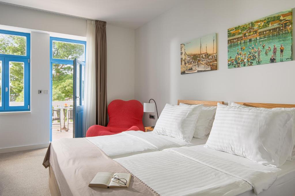 Mūsdienīga viesnīcas numura iekārtojums ar divguļamo gultu, sarkanu krēslu un zilu logu, kas ved uz terasi. Sienas rotā gleznas ar ainavām.