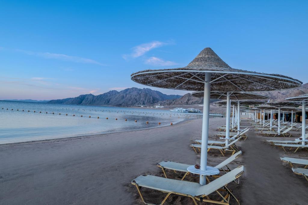 Pludmale pie Jaz Dahabeya ar saules jumtiem un kalnu fonu.