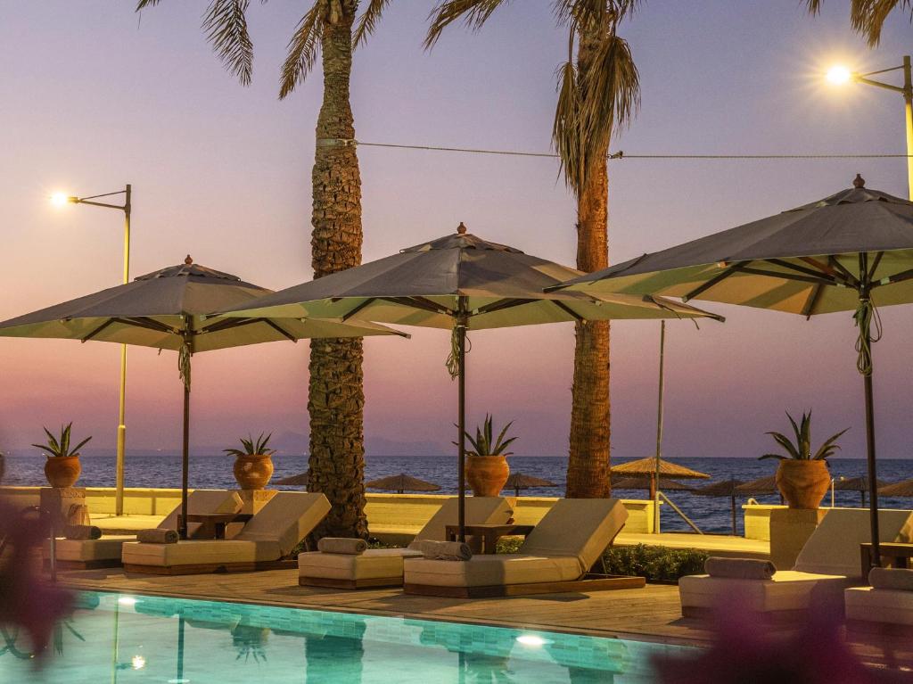 Grecotel Plaza Beach House baseins ar sauļošanās krēsliem un palmām piekrastē