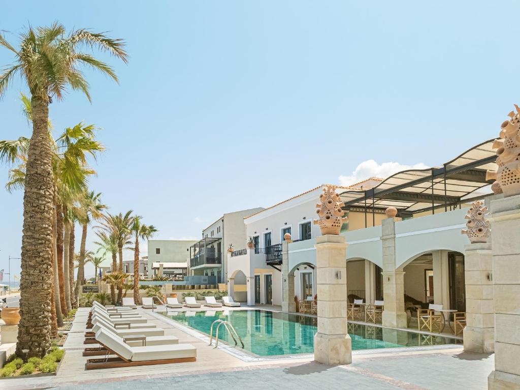 Grecotel Plaza Beach House peldbaseins ar palmām un viesnīcas ēku piekrastē.