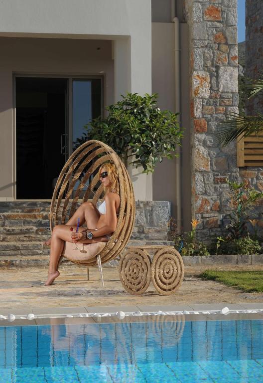 Sieviete relaksējas modernas, apaļas formas krēslā pie baseina, tērpta baltā peldkostīmā. Fons ir mūsdienīga ēka ar akmens apdari un zaļām augiem.