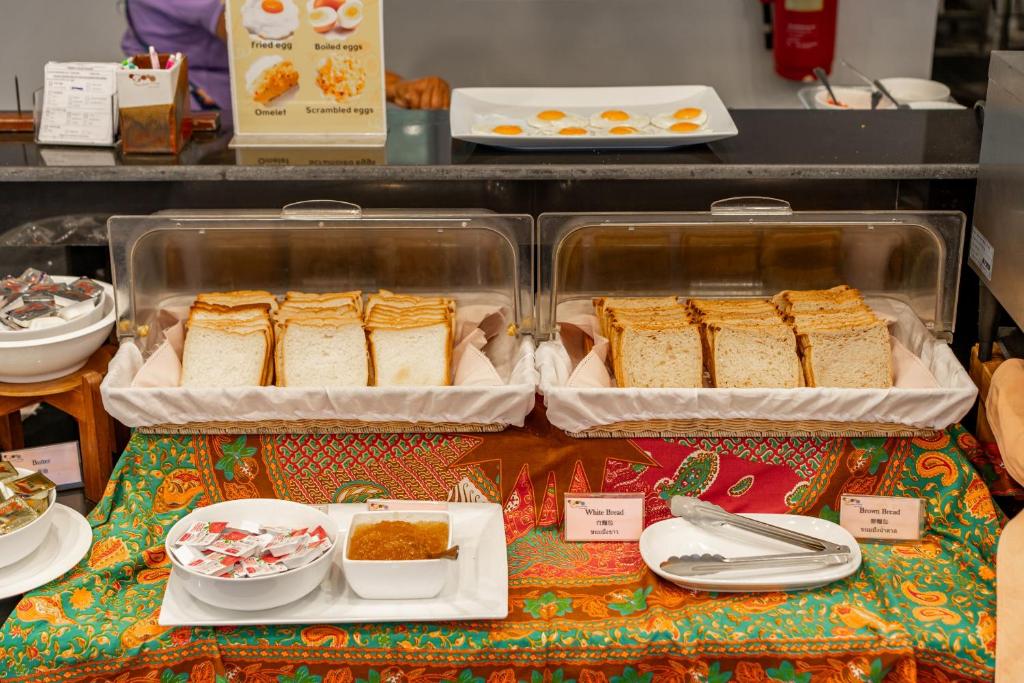 Brokastu bārs ar dažādām maizes šķēlēm, tostarp baltā un brūnā maize, kā arī dažādiem mērces iepakojumiem un ievārījumu. Fons rotāts ar krāsainu audumu.