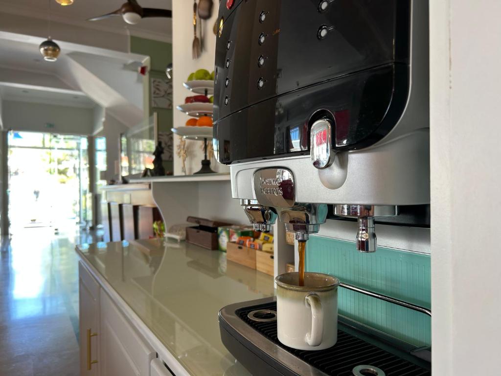 Kafijas automāts, no kura plūst kafija, novietots uz letes modernā telpā ar gaišu interjeru. Fonā redzami augļi un apgaismojums, kas rada viesmīlīgu atmosfēru.