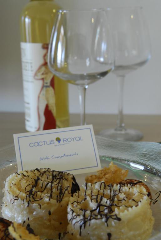 Deserts ar šokolādes pārklājumu uz šķīvja, blakus diviem glāzēm un pudeli vīna, ar zīmīti "Cactus Royal" un tekstu "With Compliments".