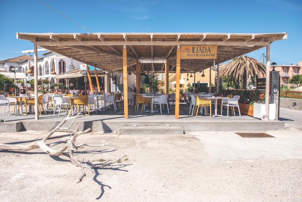 Pludmales restorāna "Ilada" ārpuses skats ar ērtām krēsliem un galdiem zem jumta, apkārtējā ainava ar zaļumiem un sauļošanās laukumiem.