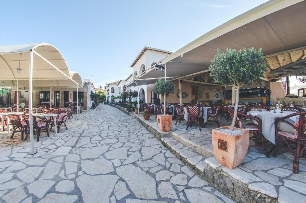 Restorāna āra zona ar akmens celiņu, galdiem un krēsliem, ko apstāda ar zaļām krūmām, radot patīkamu atmosfēru.