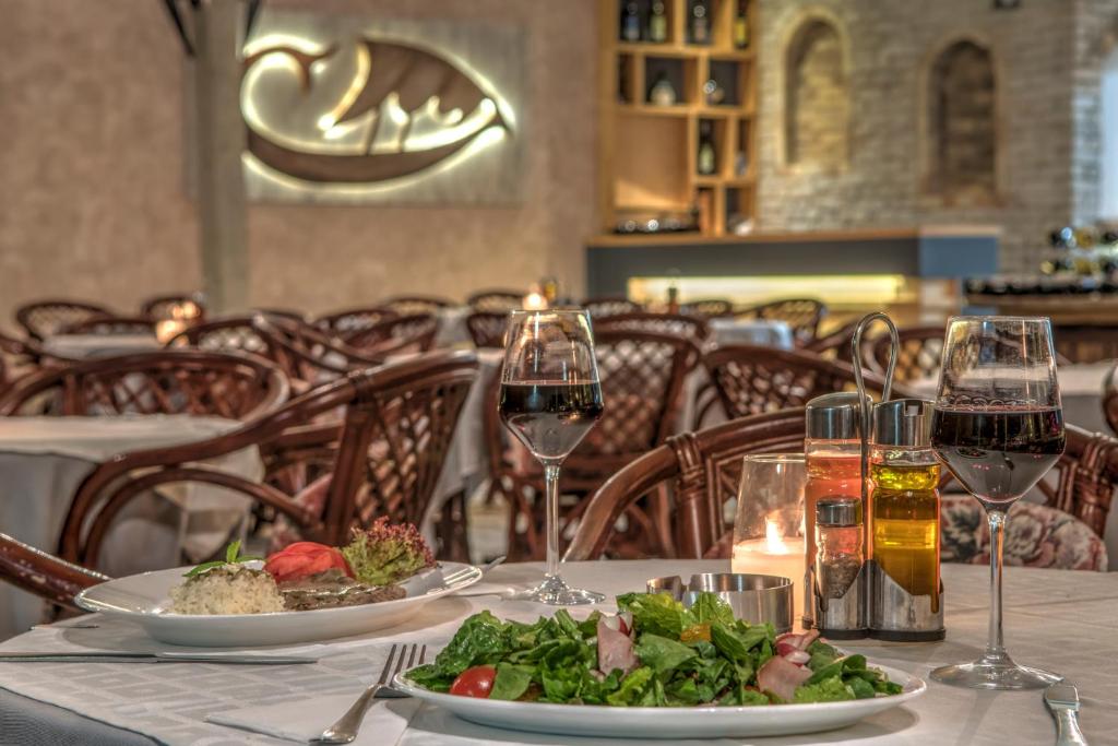 Elegantā restorāna iekārtojums ar galdiem, kas klāti ar baltām galdautiem, un izsmalcinātām maltītēm. Uz galda redzams sarkanais vīns, salātu bļoda un dažādi mērces trauki.