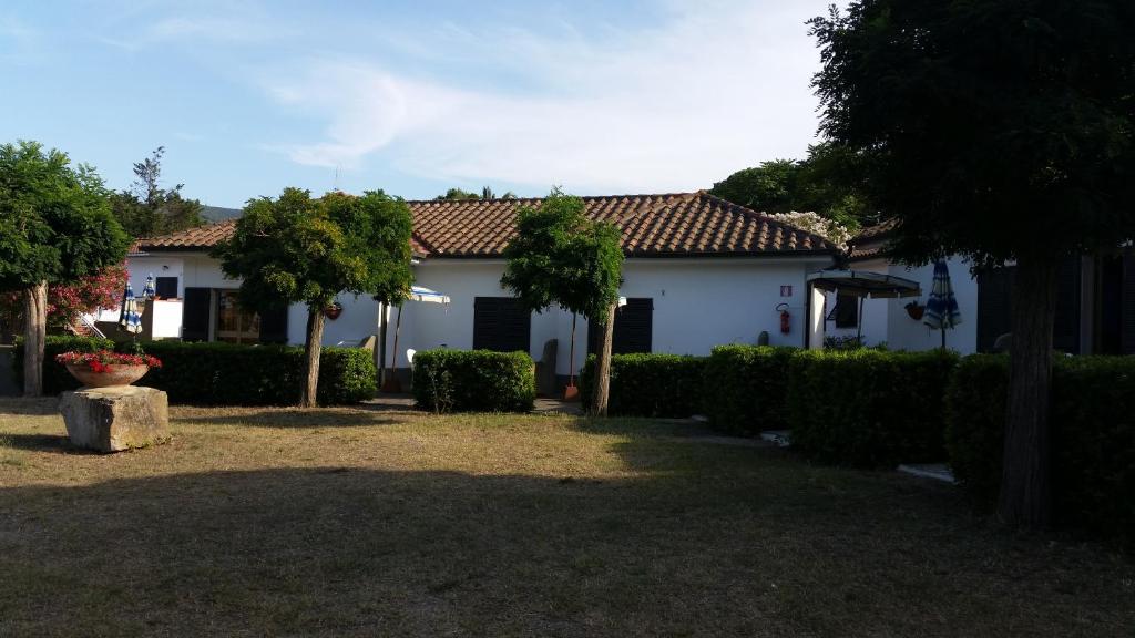 Dārza ainava ar mājīgu māju un apzaļumotu teritoriju, kurā redzami koki un krūmi. Mājai ir jumts ar flīzēm un logi ar žalūzijām, kas sniedz patīkamu atmosfēru.