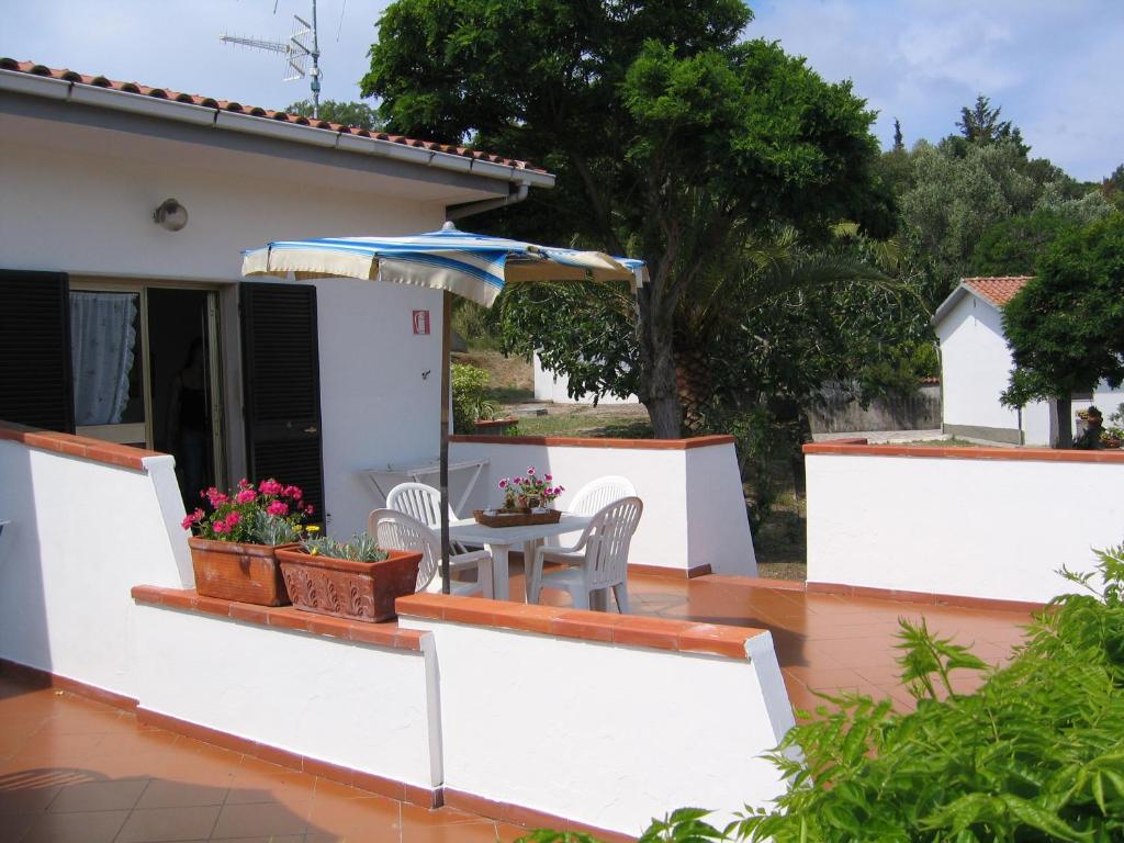 Skaista terase ar galdu un krēsliem, apstādīta ar ziediem un saulessargu, kas atrodas pie mājas. Apkārt ir zaļi koki un zāle, radot mierīgu atmosfēru.