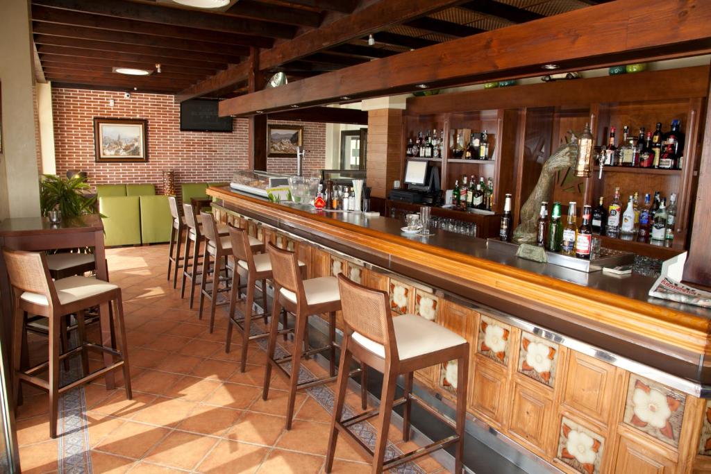 Elegants bārs ar koka apdari un flīžu grīdu, kurā ir pieejamas dažādas alkohola pudeles un sēdvietas pie bāra. Gaisma ir silta un telpa ir ērta, radot relaksējošu atmosfēru.