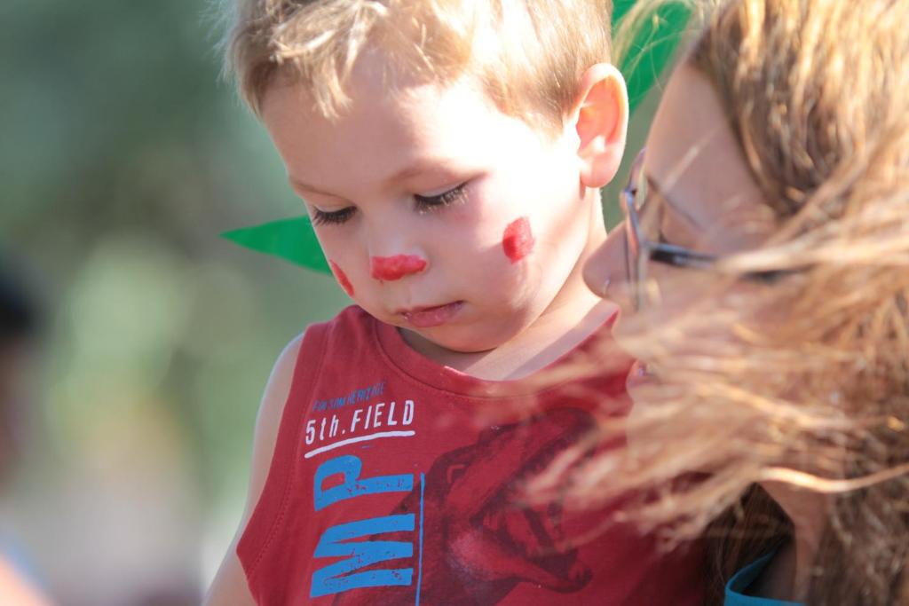 Bērns ar krāsainiem sejas zīmējumiem, kas izskatās domīgs, kamēr blakus ir pieaugušais ar brūniem matiem un brillēm. Fons ir izplūdis, radot patīkamu un mājīgu atmosfēru.
