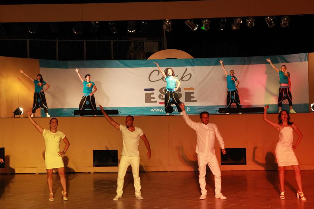 Dejotāju grupa uz skatuves izpilda energisku priekšnesumu, valkājot spilgtas drēbes un izsaka prieku ar rokām gaisā. Fons ir dekorēts ar kluba logo.