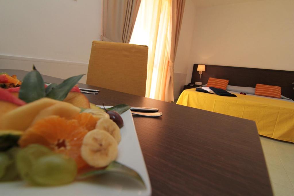 Svaigu augļu šķīvis uz galda ar viesnīcas istabu fona, kurā ir dzeltena gultas veļa un logi ar aizkariem.