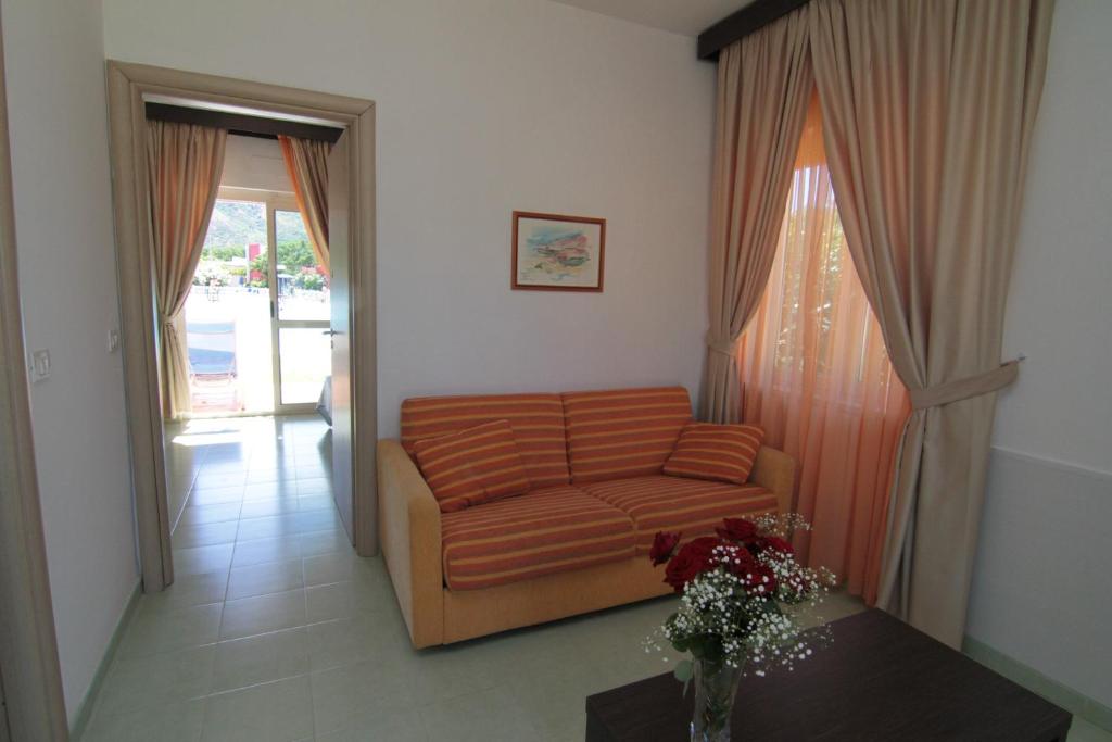 Skaista un gaiša viesistaba ar dīvānu un ziedu kompozīciju uz galda, kas rada mājīgu atmosfēru. Loga priekšā ir aizkari, kas piešķir telpai siltumu un gaismu.