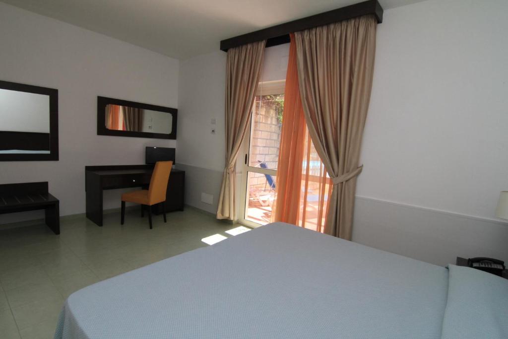 Gaiša viesnīcas numurs ar gultu, rakstāmgaldu un krēslu, blakus logam ar oranžām aizkariem. Numurs ir iekārtots ar vienkāršām mēbelēm un izgaismo to dabiska gaisma.