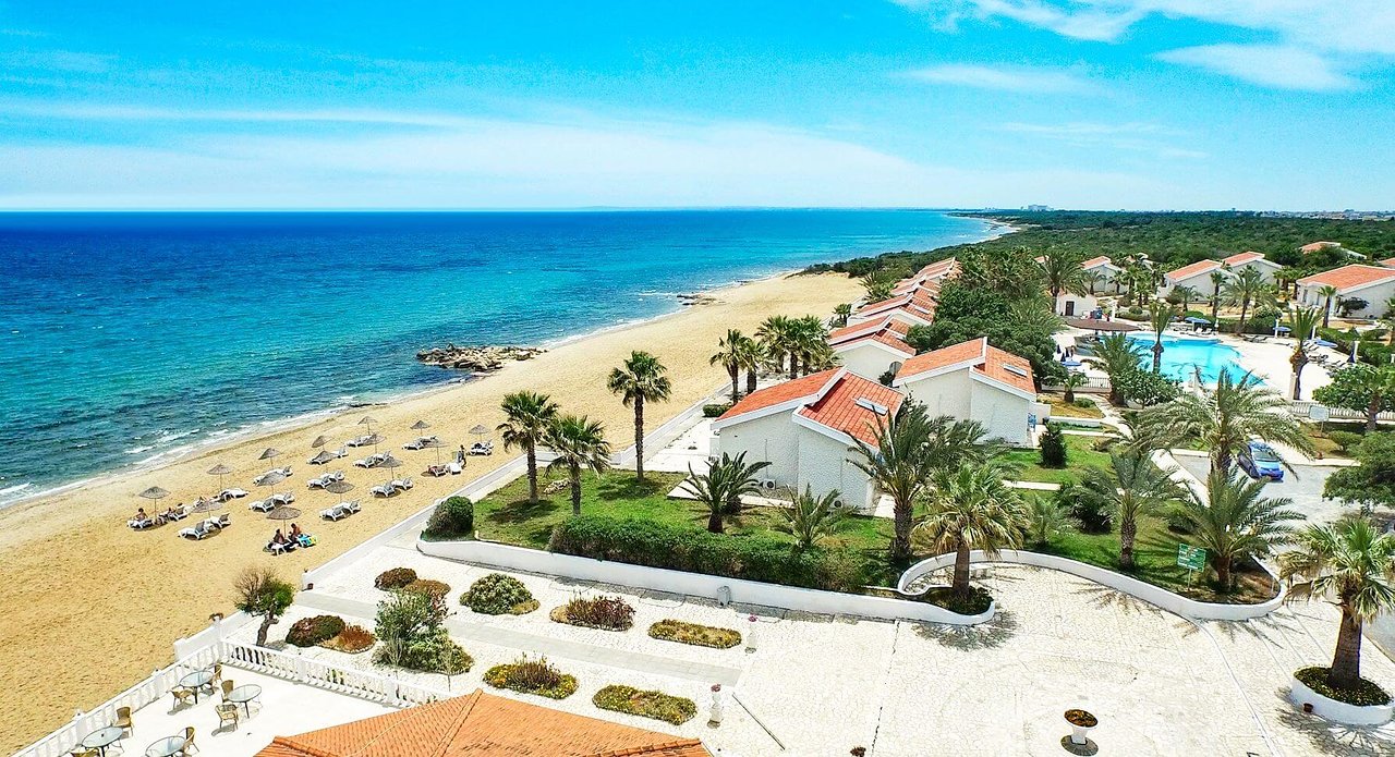 Pludmales ainava ar palmām un mājām pie jūras, kas atspoguļo atpūtas vietu un mierīgu atmosfēru. Skats uz zilo jūru un smilšu pludmali papildina kopējo ainavu. panoramic view with beach and pools