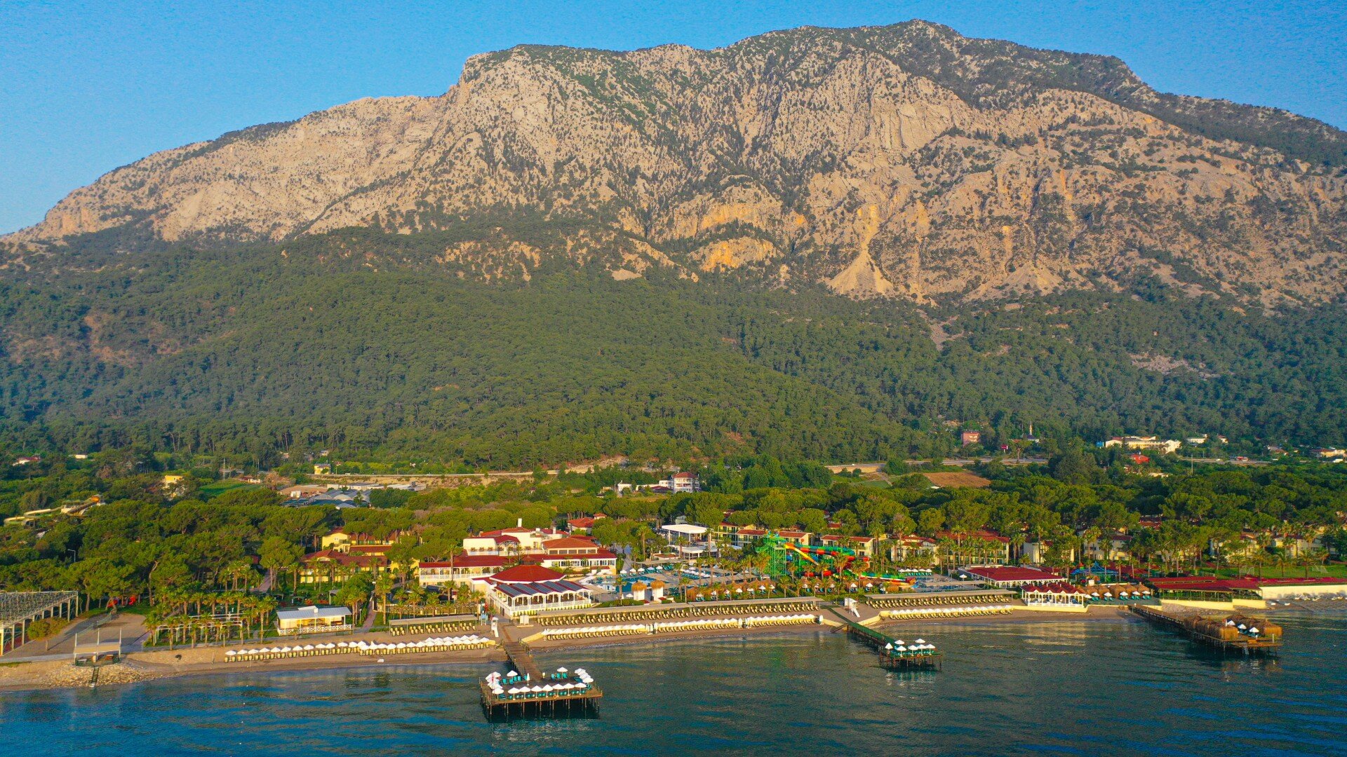 Krāšņs kalnu skats ar zaļām meža ainavām un pludmales kūrortu pie ūdens, atspoguļojot mierīgu vasaras atmosfēru. panoramic view with beach and pools