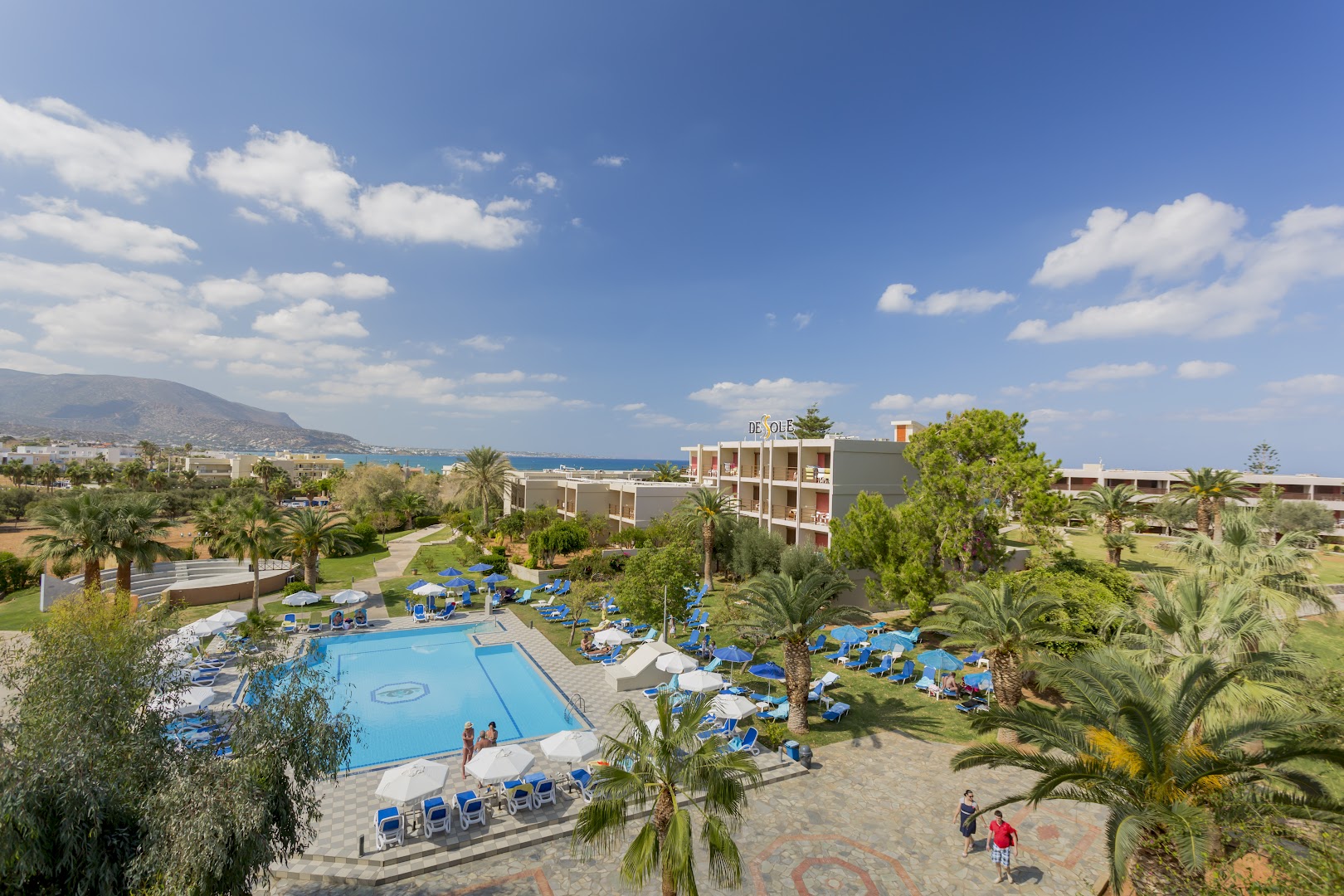 Panorāmas skats uz Dessole Malia Beach āra baseinu, sauļošanās zonu ar saulessargiem un viesnīcas korpusiem pie jūras Krētā