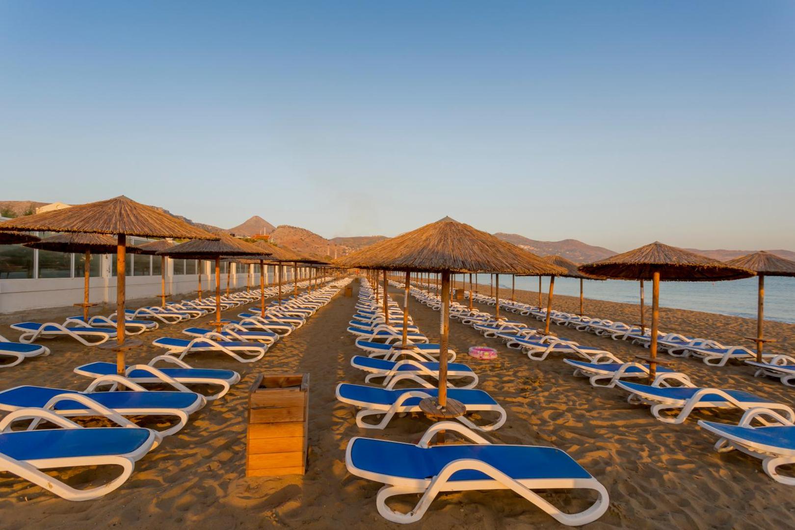 Tuvplāns ar salmu saulessargiem un ziliem sauļošanās krēsliem Dessole Malia Beach pludmalē saulrieta vai saullēkta gaismā