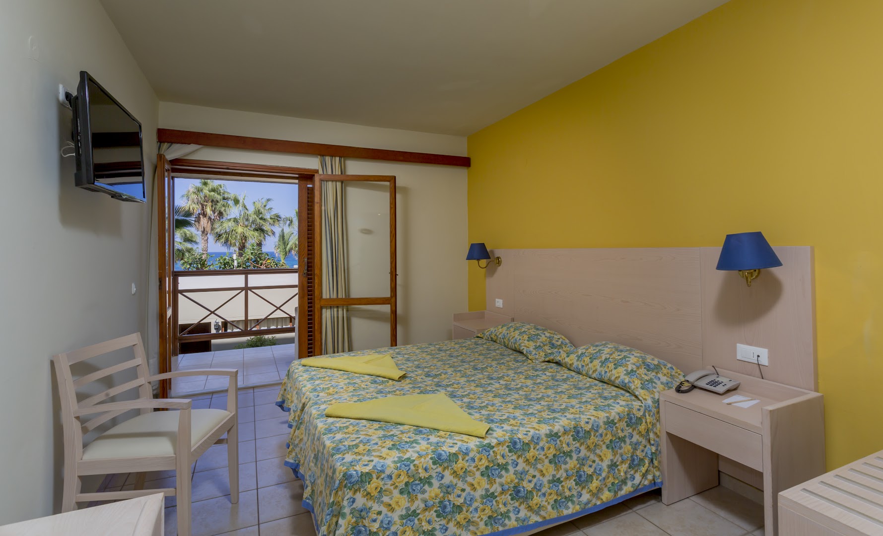 Dessole Malia Beach numurs ar dzeltenu akcenta sienu, ziedu raksta gultas pārklāju un balkonu ar skatu uz palmu dārzu