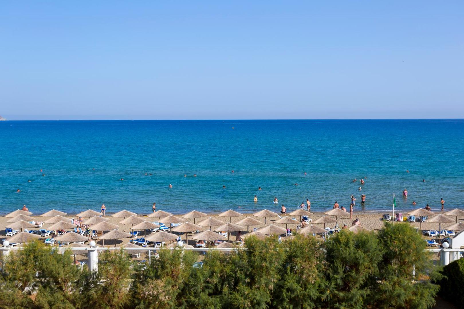 Dessole Malia Beach viesnīcas pludmale Krētā ar salmu saulessargiem un zilu jūru — skats no augšas uz atpūtniekiem un piekrastes joslu