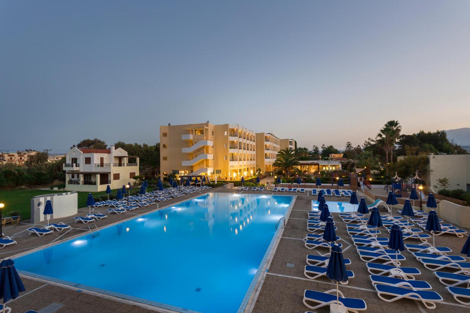 Dessole Malia Beach viesnīcas galvenais baseins vakarā Malijā, Krētā — apgaismota teritorija ar sauļošanās krēsliem un viesnīcas korpusiem fonā