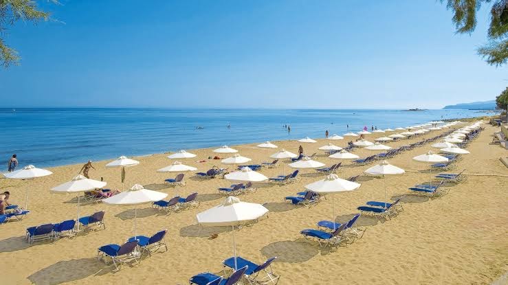 Gara smilšu pludmales josla pie Dessole Malia Beach viesnīcas ar rindās sakārtotiem baltiem saulessargiem un ziliem sauļošanās krēsliem
