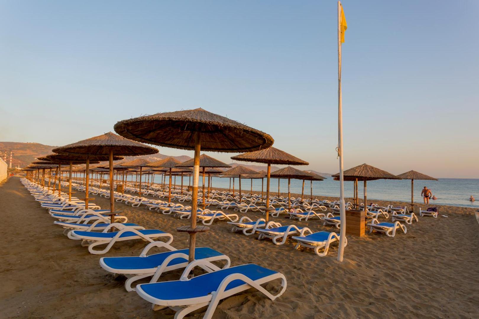Rindas ar saulessargiem un guļamkrēsliem Dessole Malia Beach pludmalē, fonā jūra un kalnu silueti vakara saulē