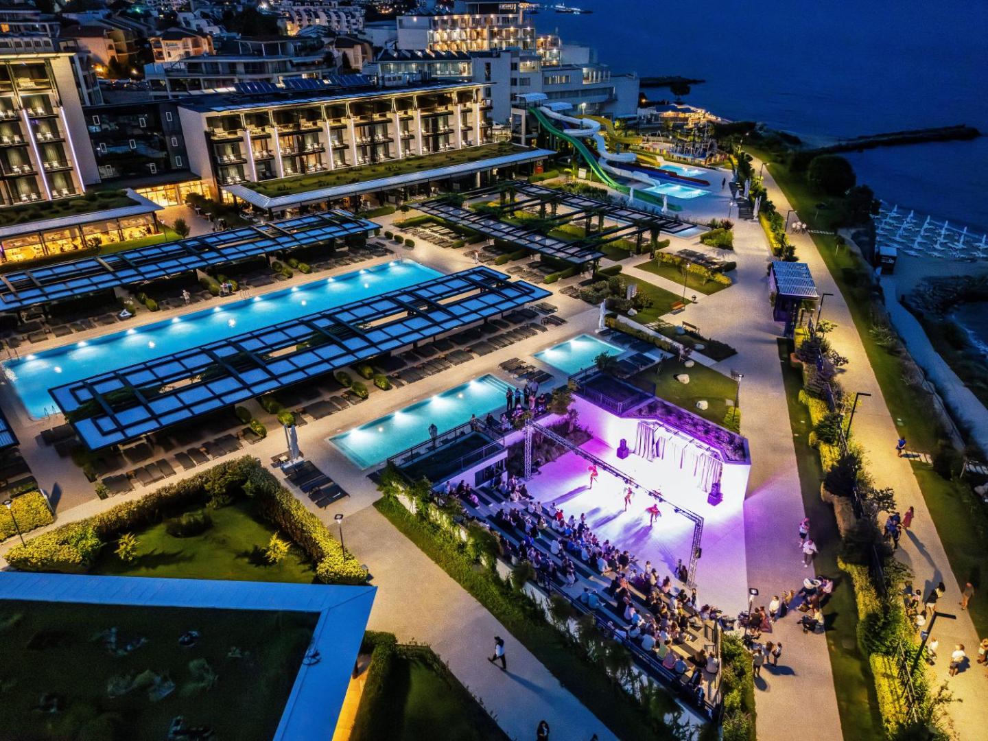 Voya Beach Resort vakara apgaismojums pie baseiniem un āra skatuves — izgaismota teritorija, atpūtas zonas un viesnīcas korpusi naktī