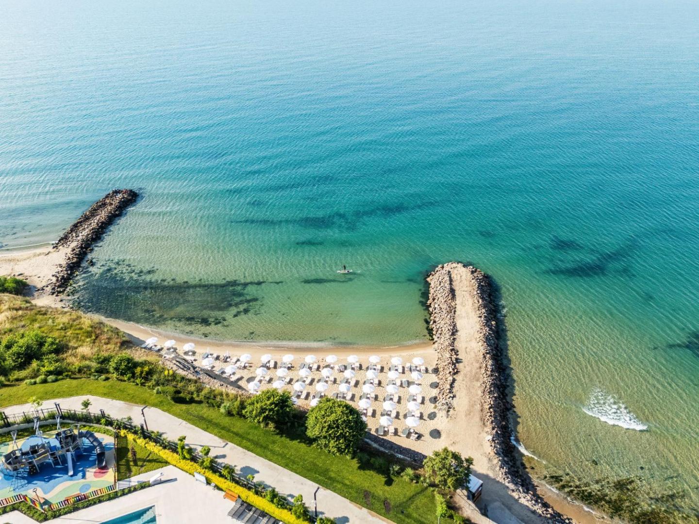 Aerofoto skats uz Voya Beach Resort privāto pludmali ar saulessargiem, akmens moliem un tirkīzzilu jūras ūdeni.