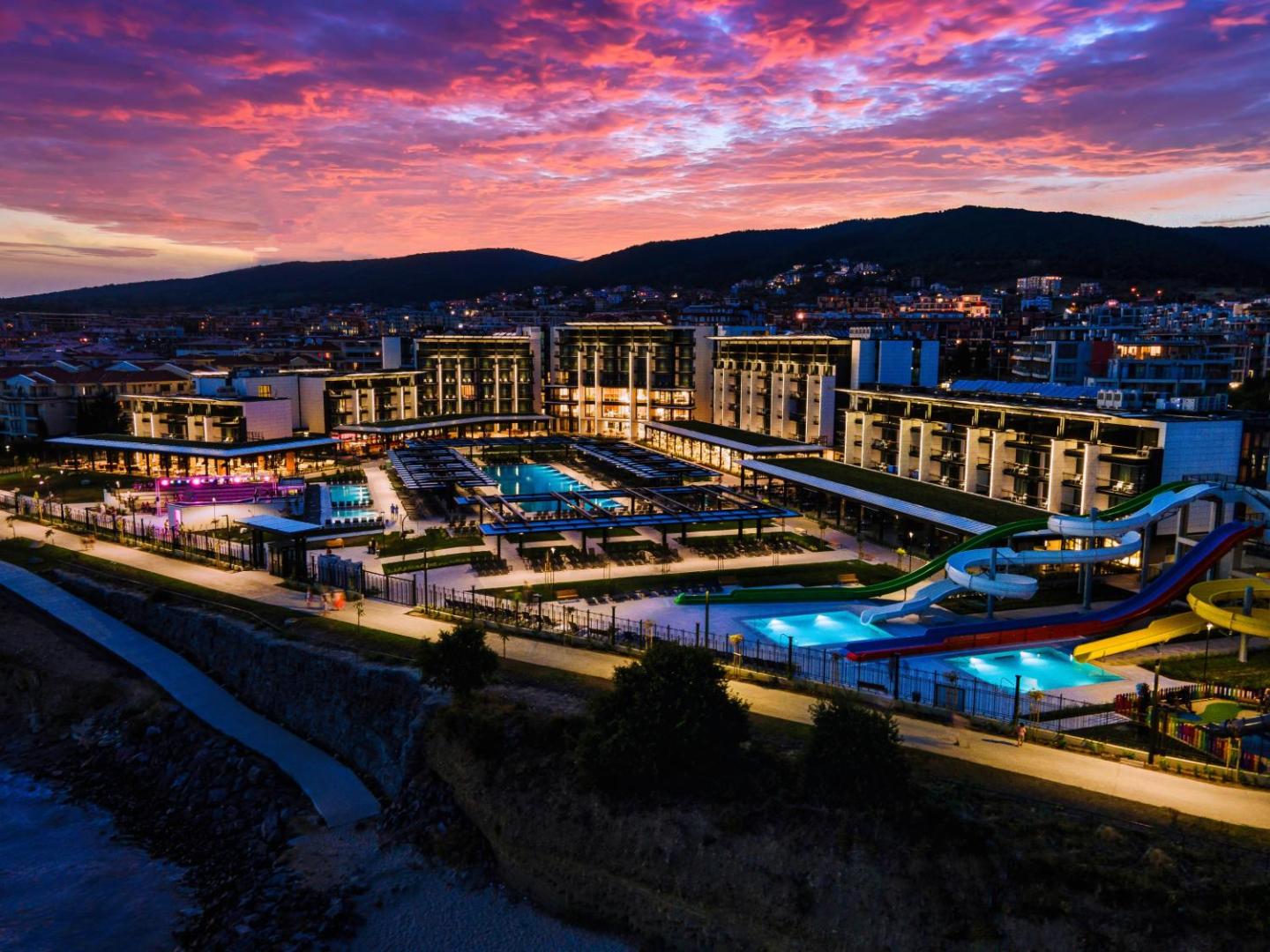 Viesnīca Voya Beach Resort krāšņa saulrieta laikā — debesis purpura toņos virs modernās arhitektūras, baseiniem un akvaparka Sveti Vlasā