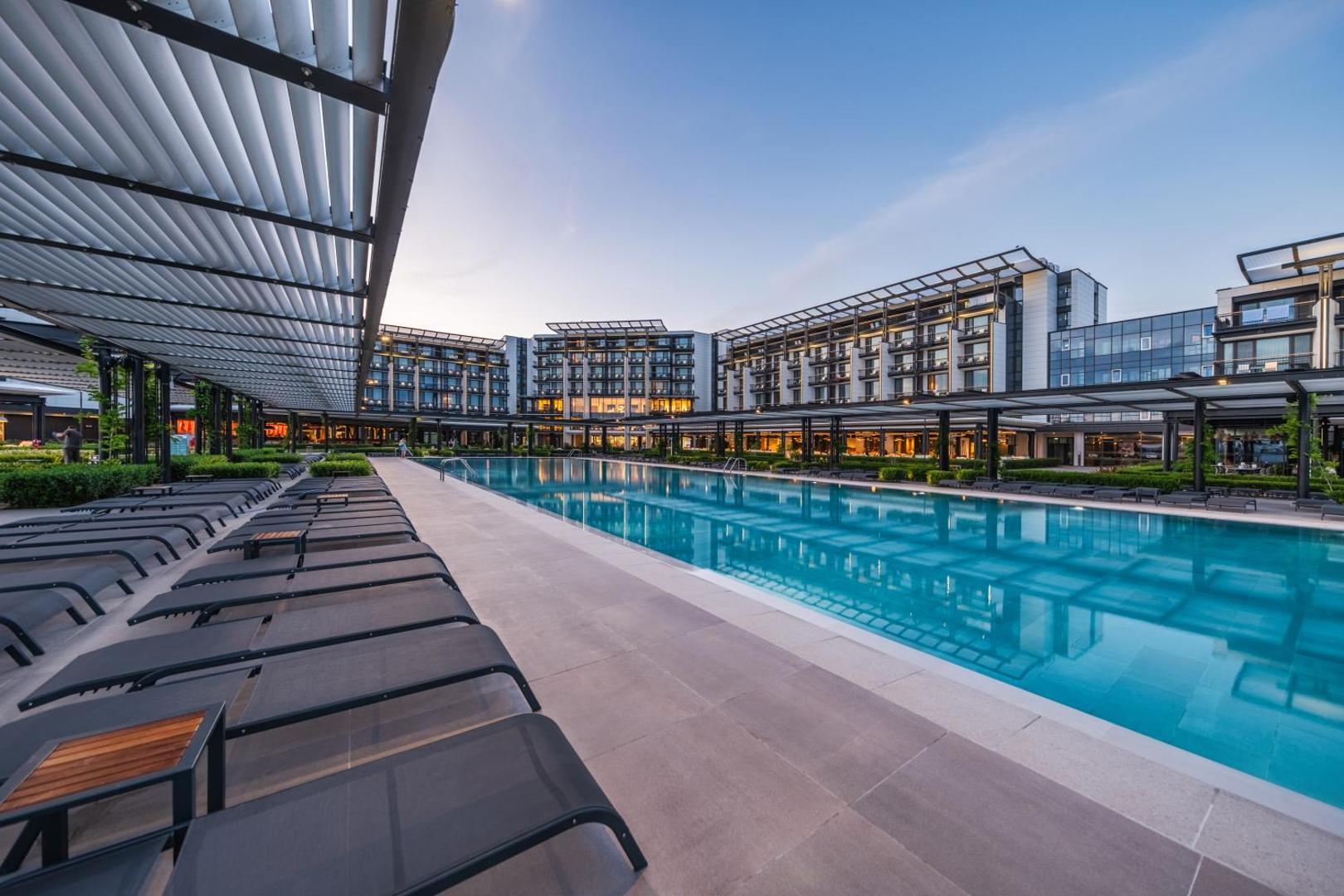 Voya Beach Resort baseina zona tuvplānā ar ērtiem zviļņiem zem nojumēm un skatu uz viesnīcas moderno fasādi vakarā