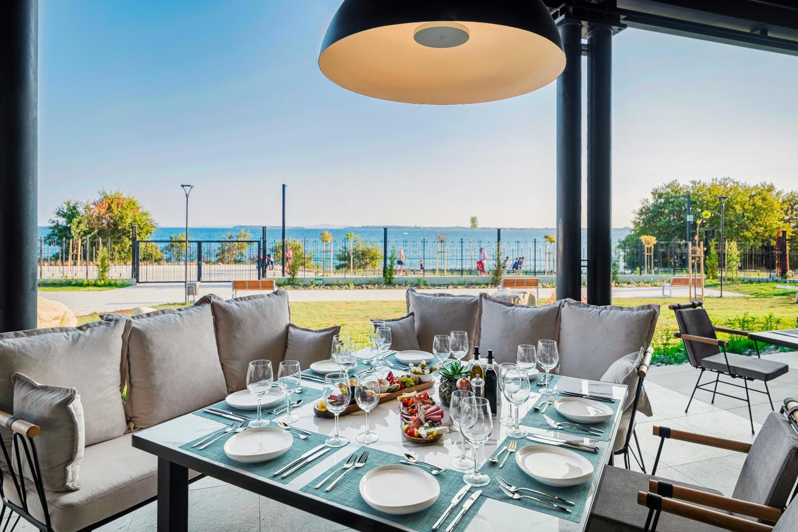 Voya Beach Resort restorāna terase ar klātu galdu, ērtiem dīvāniem un skatu uz jūras piekrasti saulainā dienā.