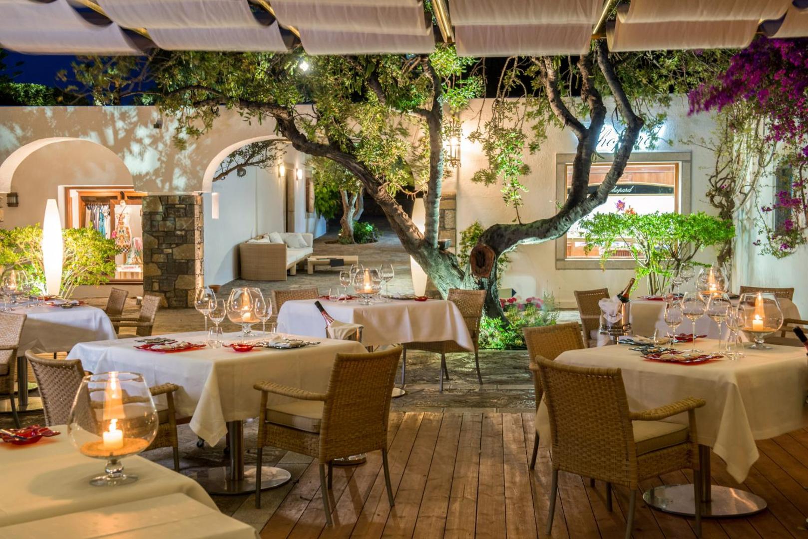 Elounda Beach Hotel And Villas restorāns vakarā ar klātiem galdiem, koka grīdu un atmosfērisku apgaismojumu dārzā