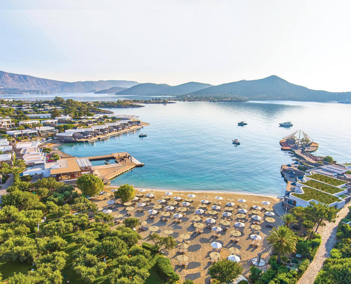 Elounda Beach Hotel And Villas skats no augšas uz privāto smilšu pludmali, saulessargiem un zilo līci Krētā, Grieķijā.